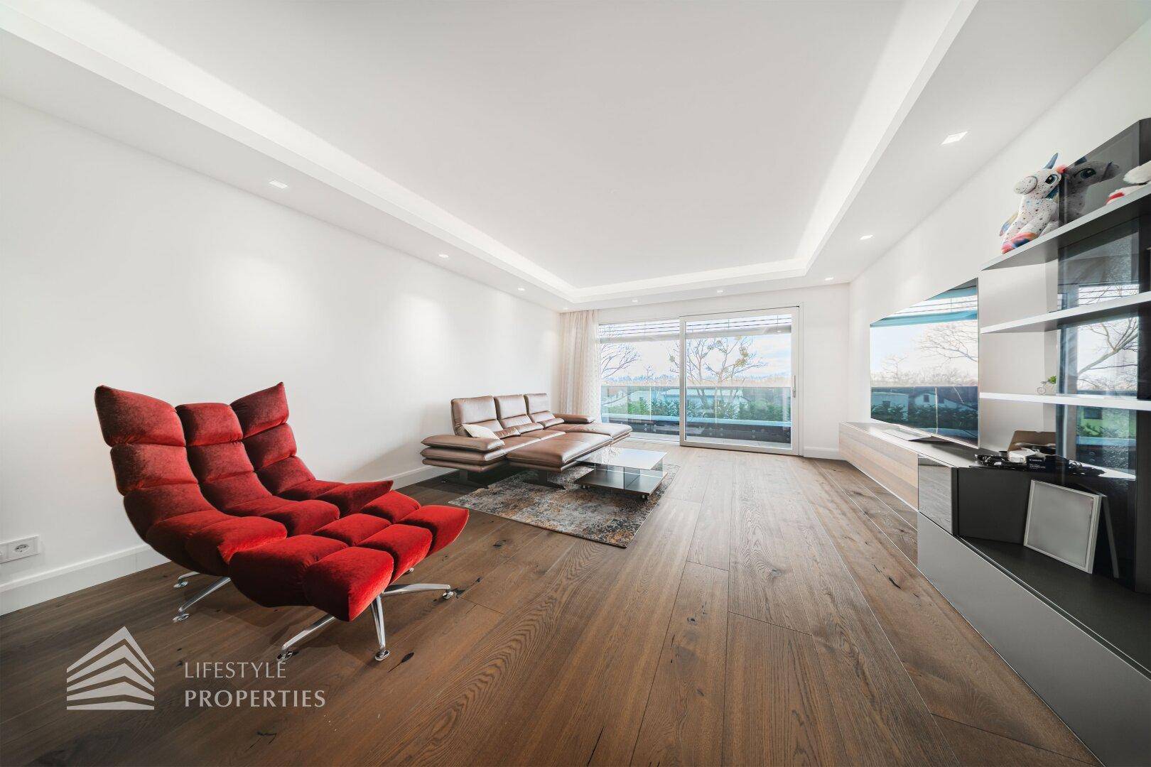 Geräumiges Wohnzimmer mit Holzboden, rotem Sessel und großem Fenster mit Blick nach draußen.
