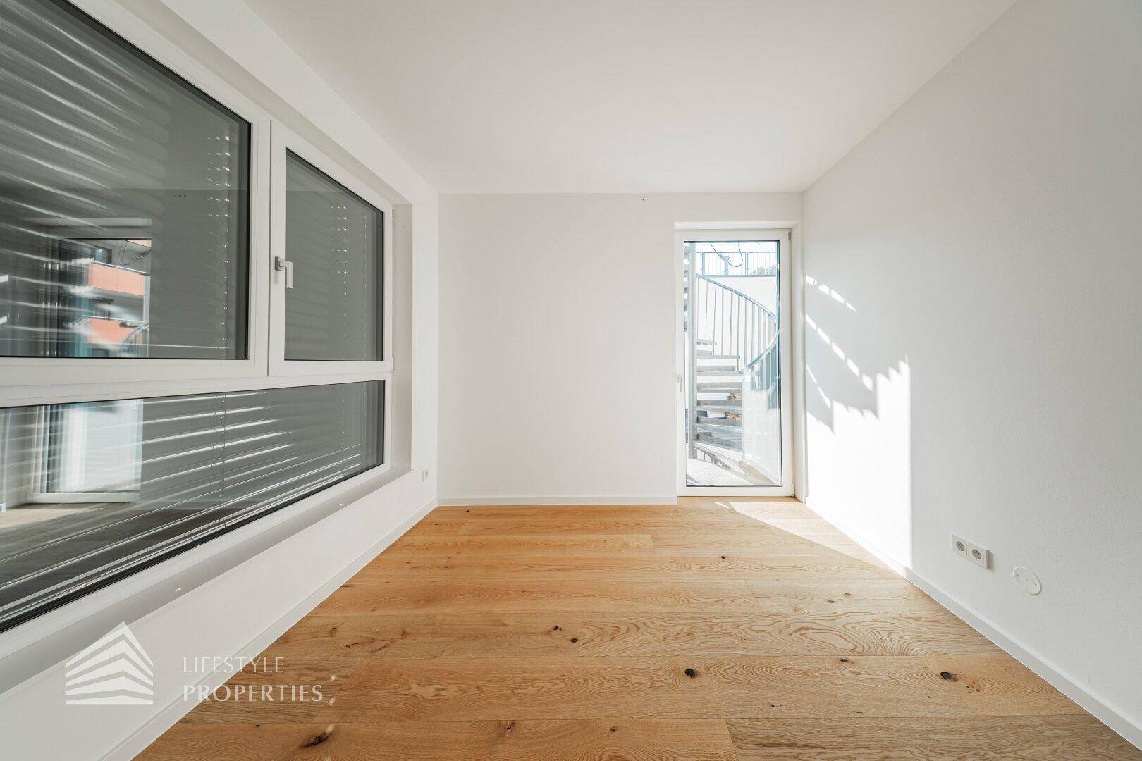 Heller Raum mit Holzboden, großen Fenstern und Zugang zu einer Außentreppe.