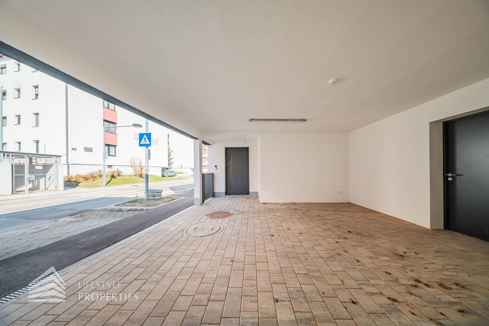 Überdachter Parkplatz mit Pflastersteinen und Zugang zum Gebäude, an einer ruhigen Straße gelegen.