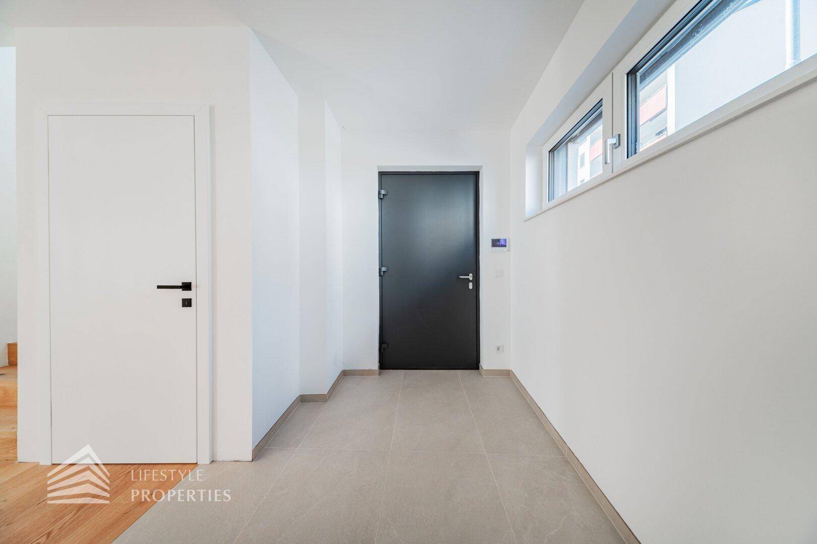 Heller Flur mit Fliesenboden, weißer Tür und dunkler Eingangstür, beleuchtet durch ein Fenster.