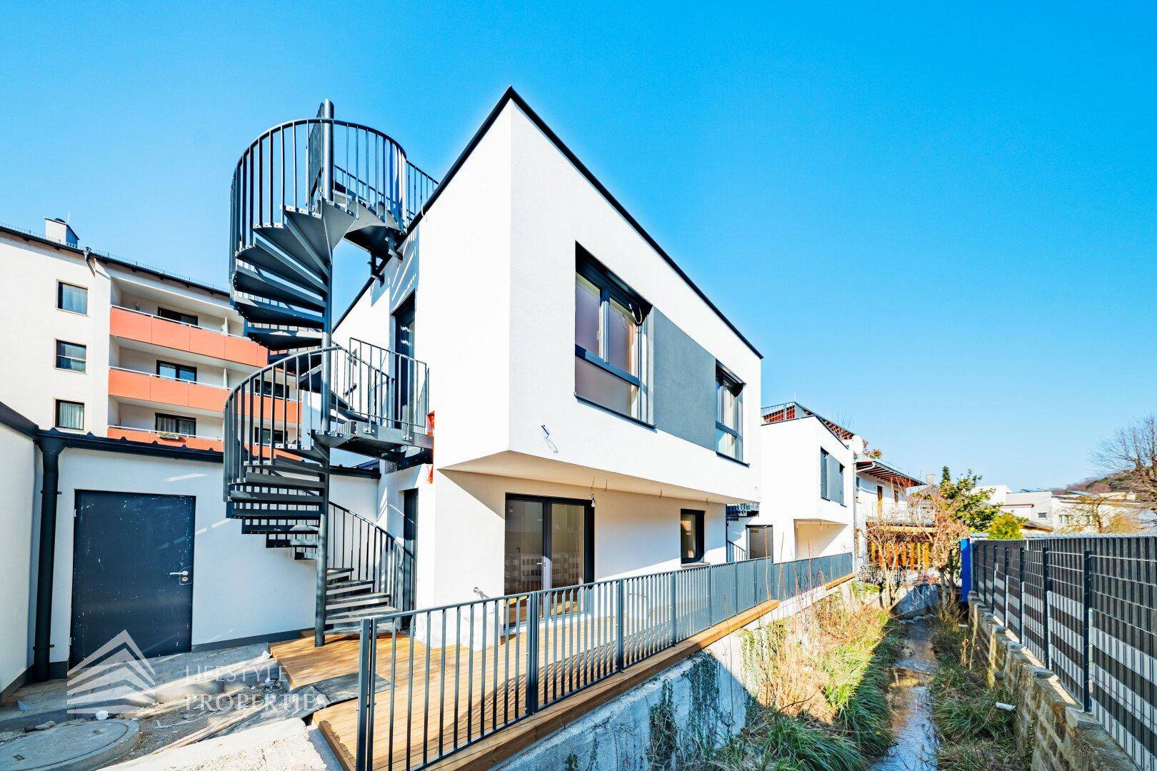 Moderne weiße Fassade mit schwarzer Wendeltreppe und angrenzendem Bachlauf, in einer grünen Umgebung.