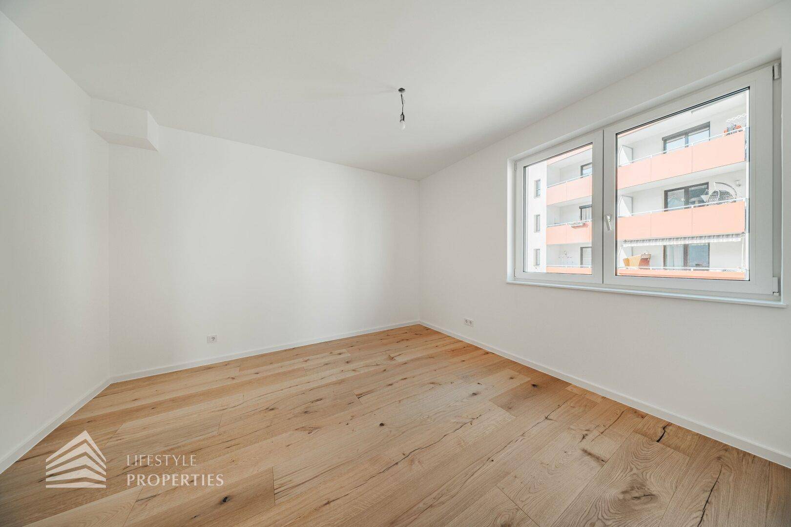Leeres Schlafzimmer mit Holzdielenboden und großem Fenster, das viel Licht spendet.