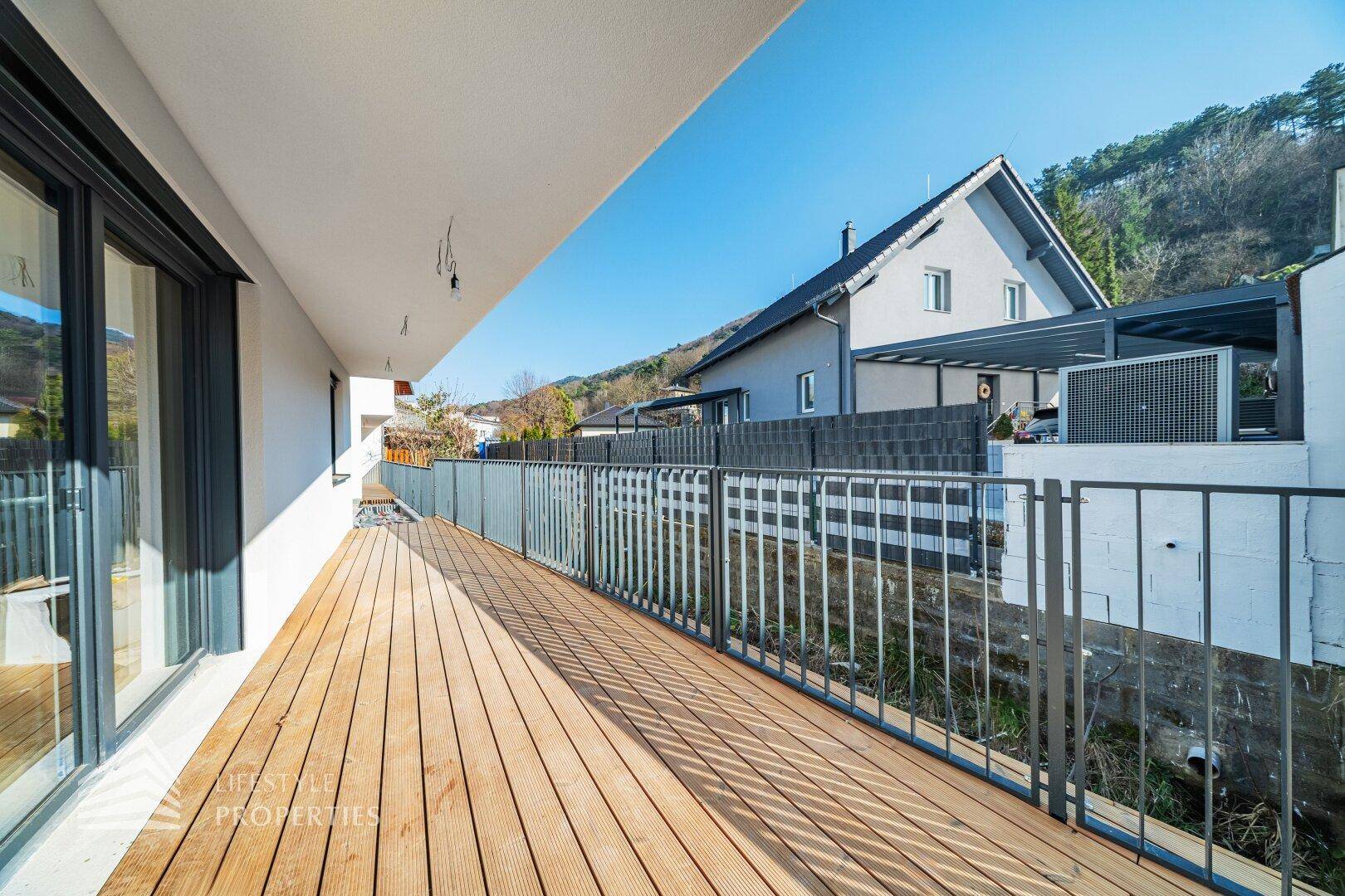 Langer, sonniger Holzbalkon mit Metallgeländer und Blick auf die umliegenden Häuser und Hügel.