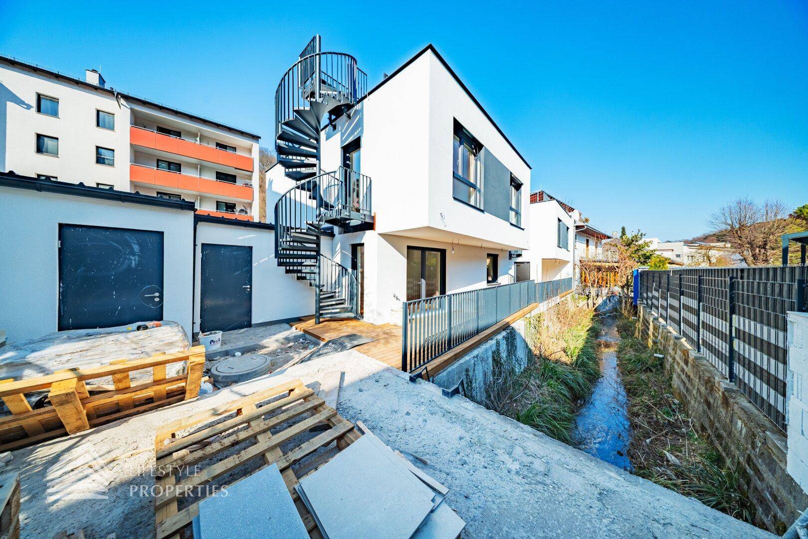 Außenansicht des modernen Neubaus mit Wendeltreppe und angrenzendem Bach, noch im Bauzustand.