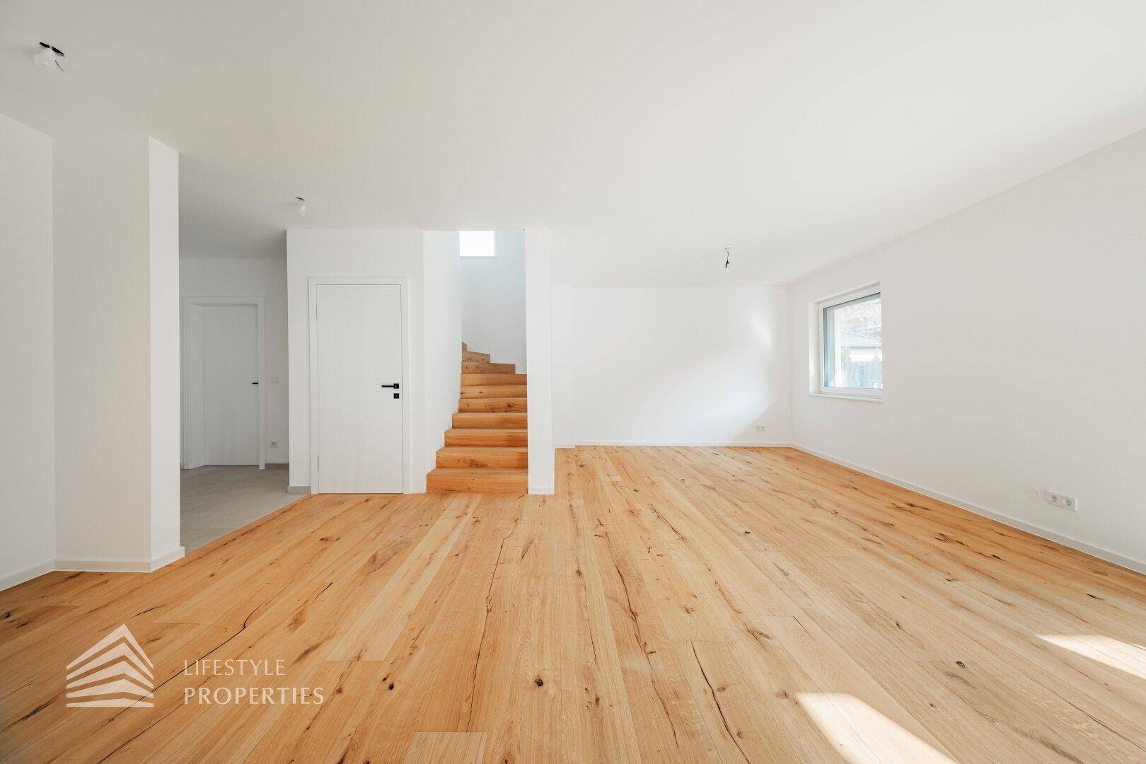 Geräumiges, helles Zimmer mit Holzboden und einer modernen Holztreppe, die in die obere Etage führt.