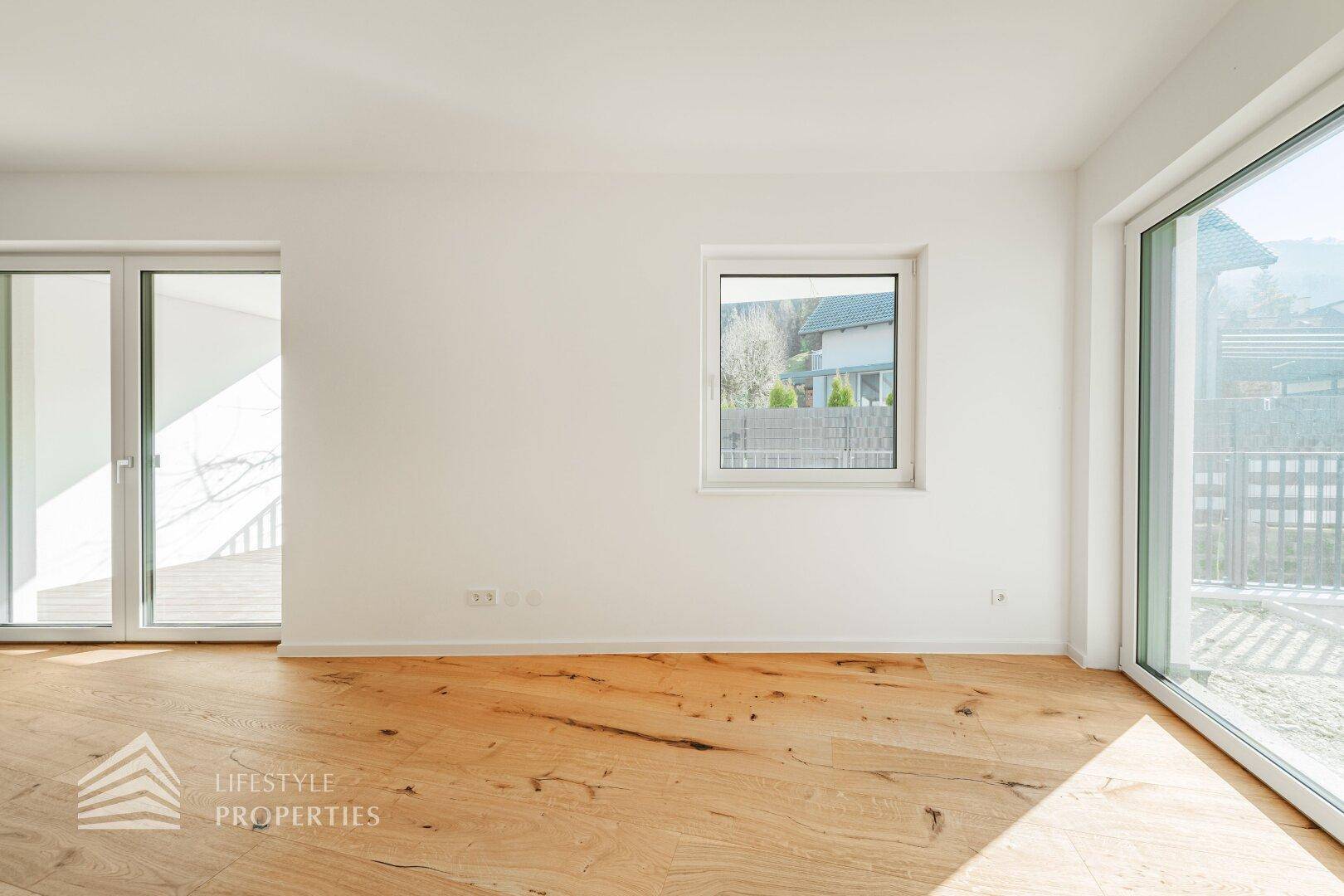 Leeres Wohnzimmer mit Holzboden und mehreren Fenstern, die einen Blick ins Grüne ermöglichen.