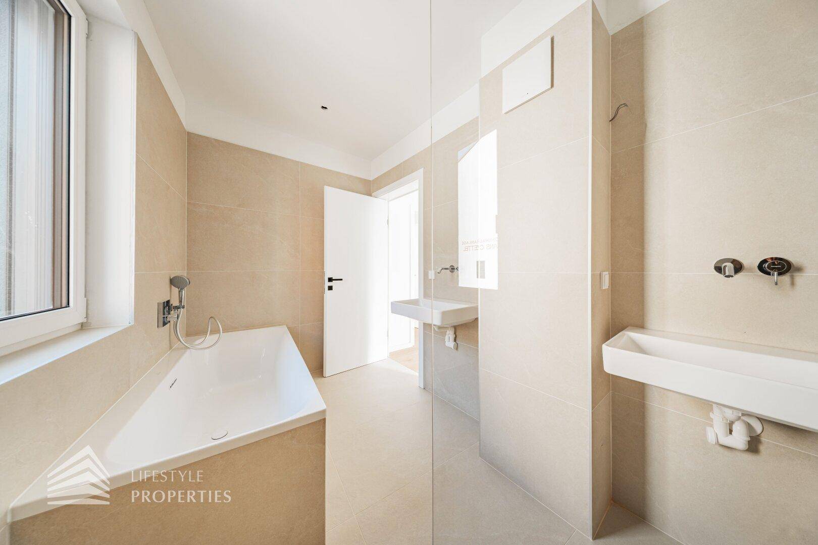 Helles Badezimmer mit beigefarbenen Fliesen, Badewanne, Dusche und zwei modernen Waschbecken.