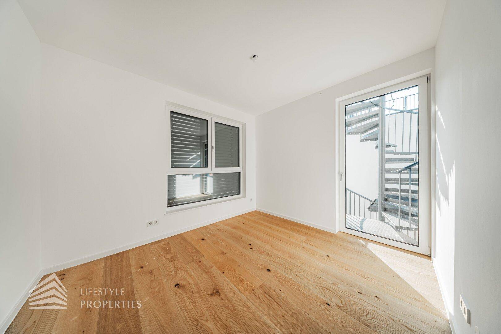 Heller Raum mit Holzboden, Fenster und Zugang zu einer Außentreppe.