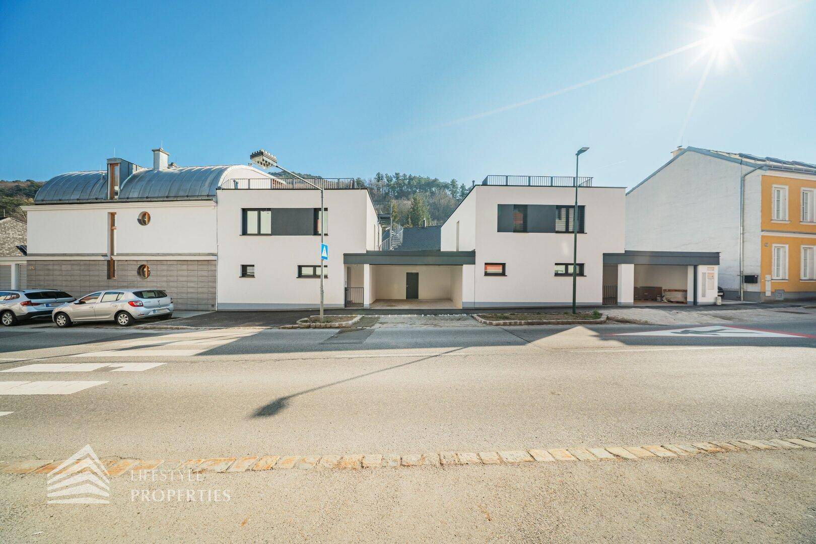 Moderne weiße Gebäude mit Carports an einer Straße, umgeben von grünen Hügeln.