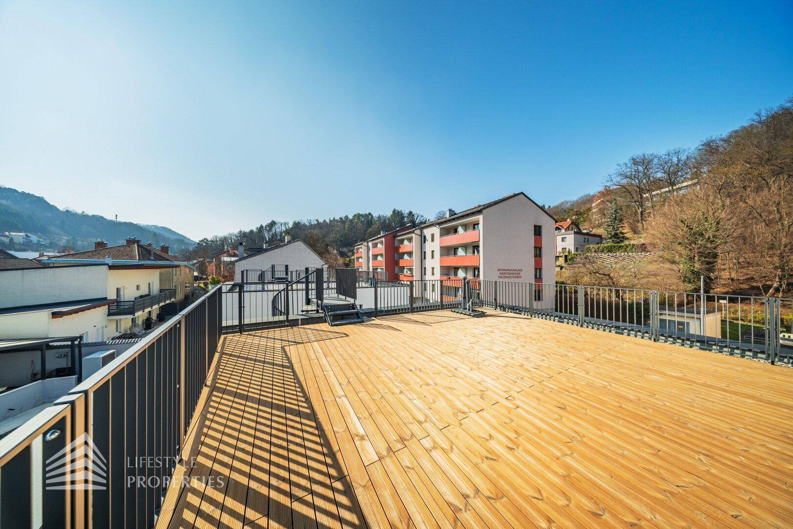 Weitläufige Dachterrasse mit Holzdielen und Blick auf benachbarte Gebäude und grüne Hügel.