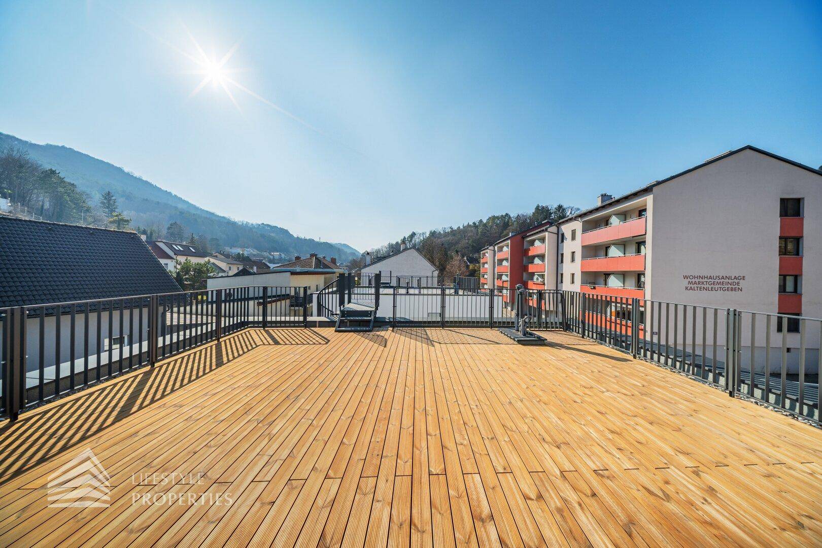 Weitläufige Dachterrasse mit Holzdielen und Blick auf benachbarte Gebäude und grüne Hügel.