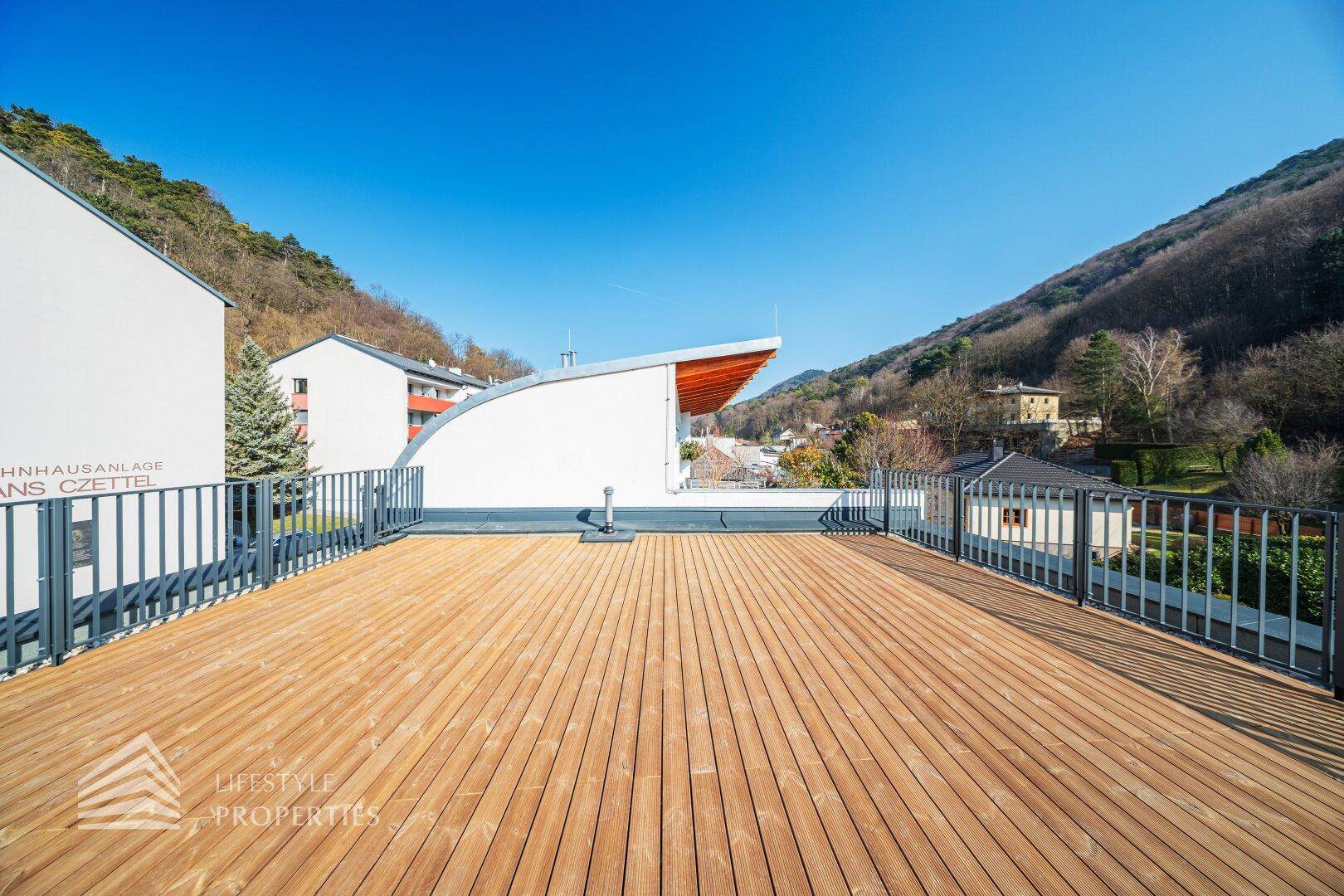 Große Dachterrasse mit Holzboden und Blick auf ein modernes Gebäude und die umliegenden Hügel.