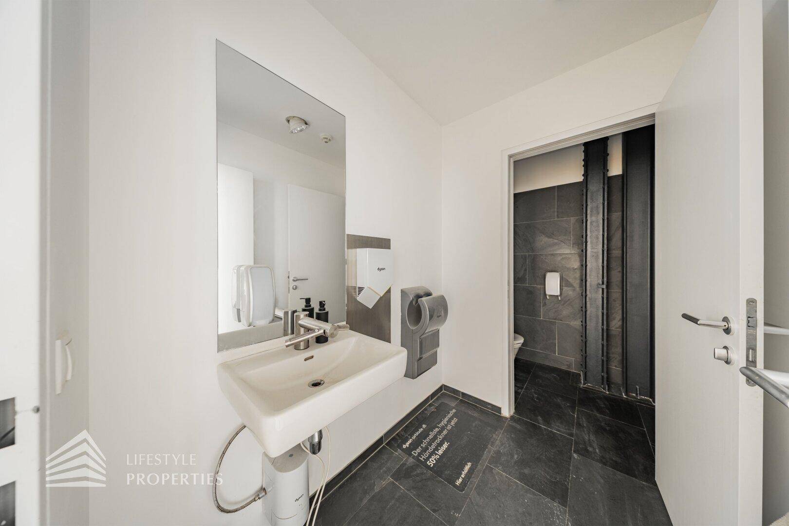 Modernes Badezimmer mit Waschbecken, Spiegel und dunklen Fliesen im Duschbereich.