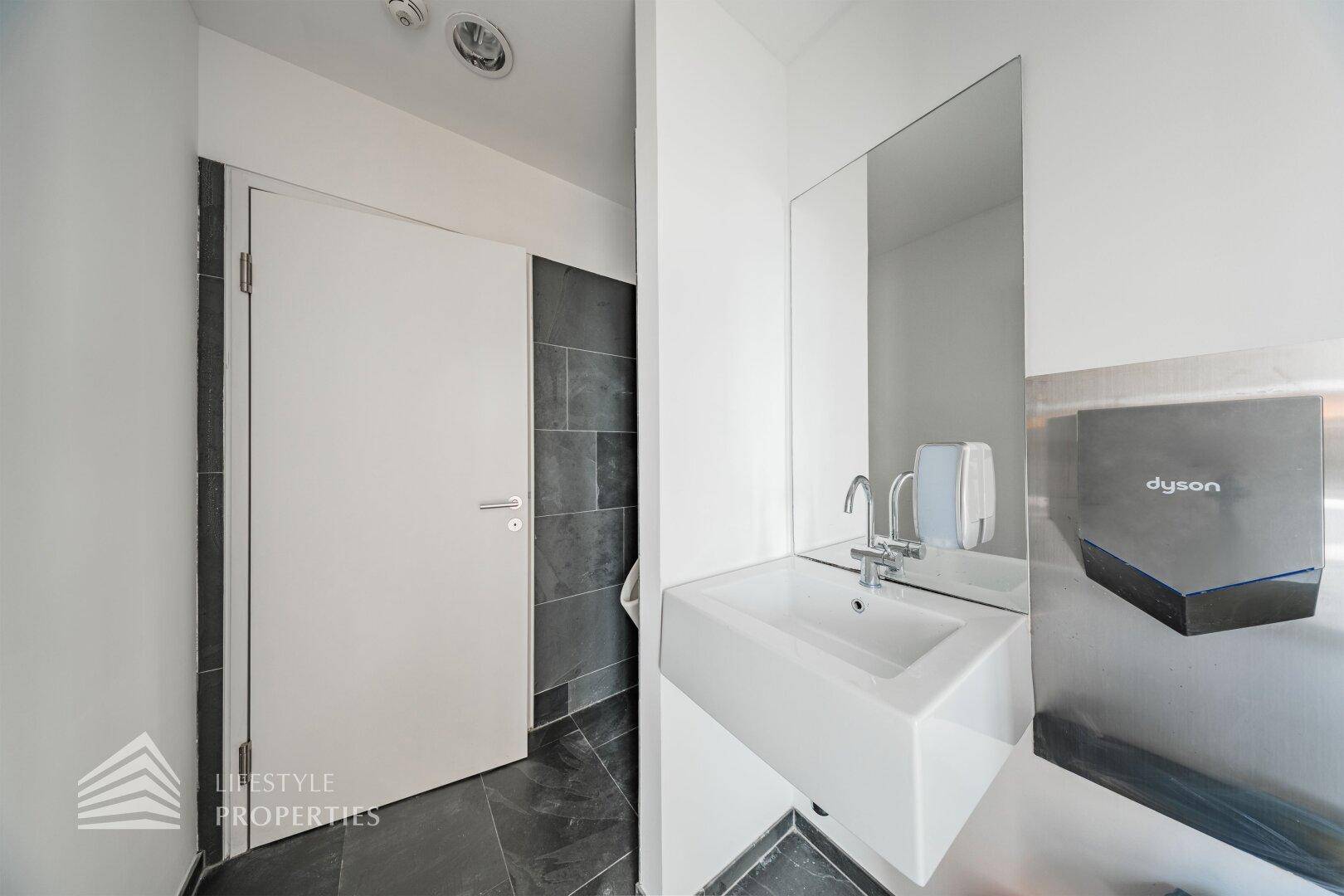 Modernes Badezimmer mit Waschbecken, Spiegel und Handtrockner, gefliest mit dunklen Steinplatten.