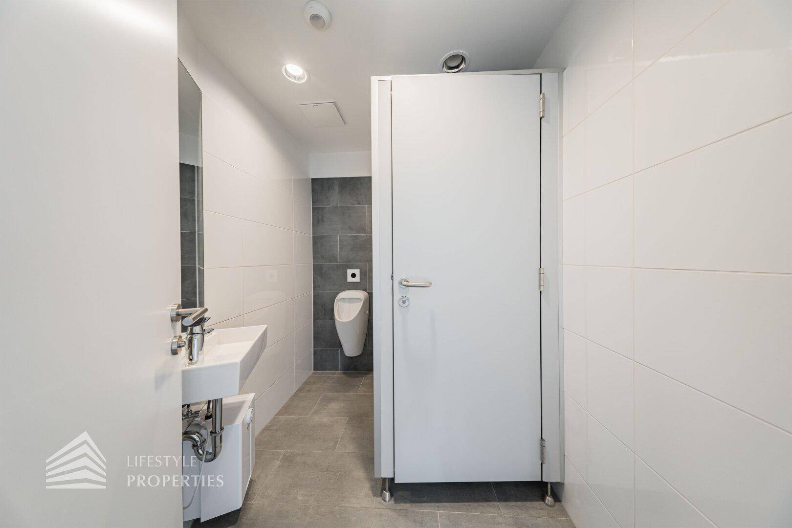 Moderner Toilettenbereich mit Waschbecken, Urinal und weiß-grauen Fliesen, sauber und funktional.