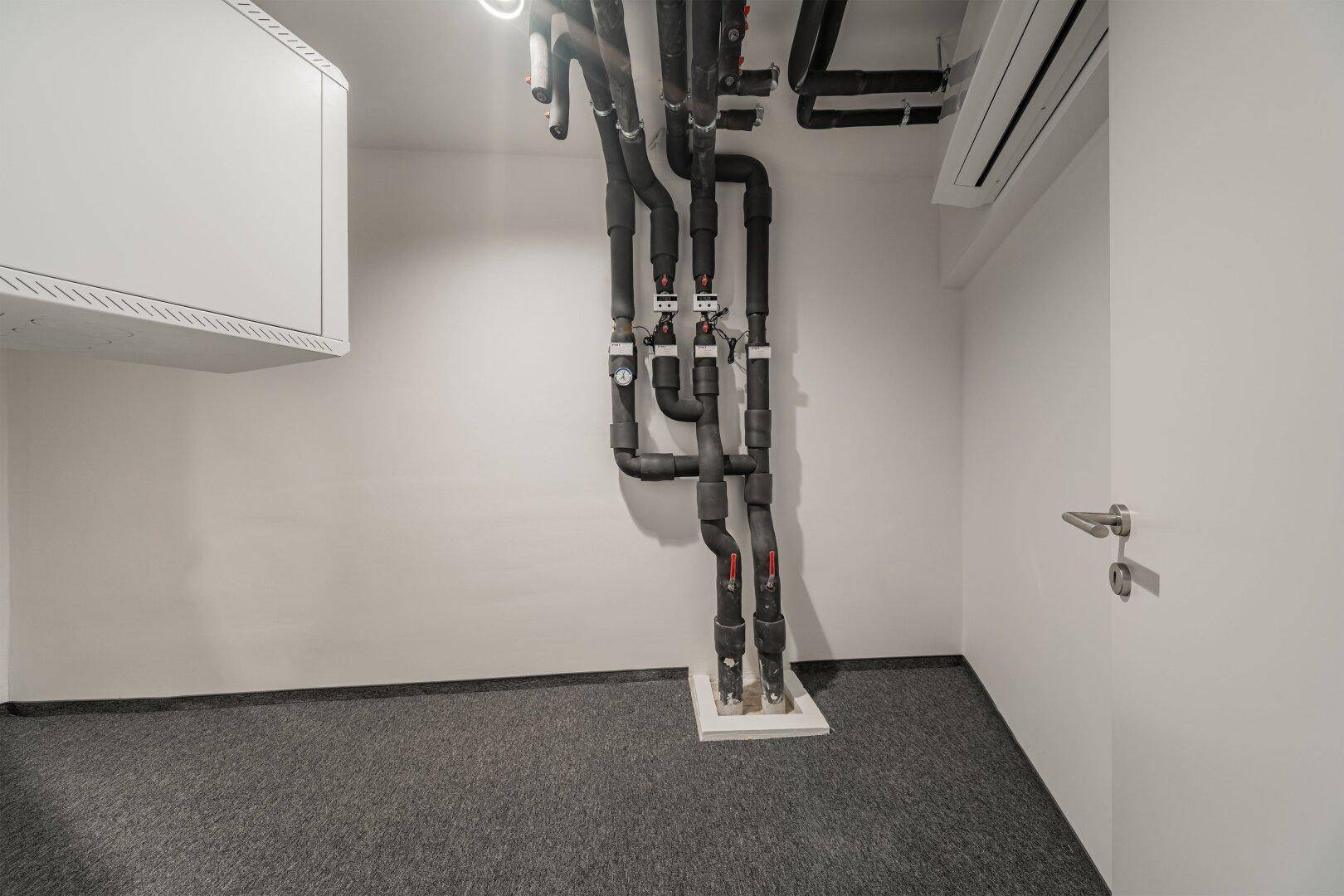 Technikraum mit sichtbaren Rohren und Leitungen an der Decke und Klimaanlage.