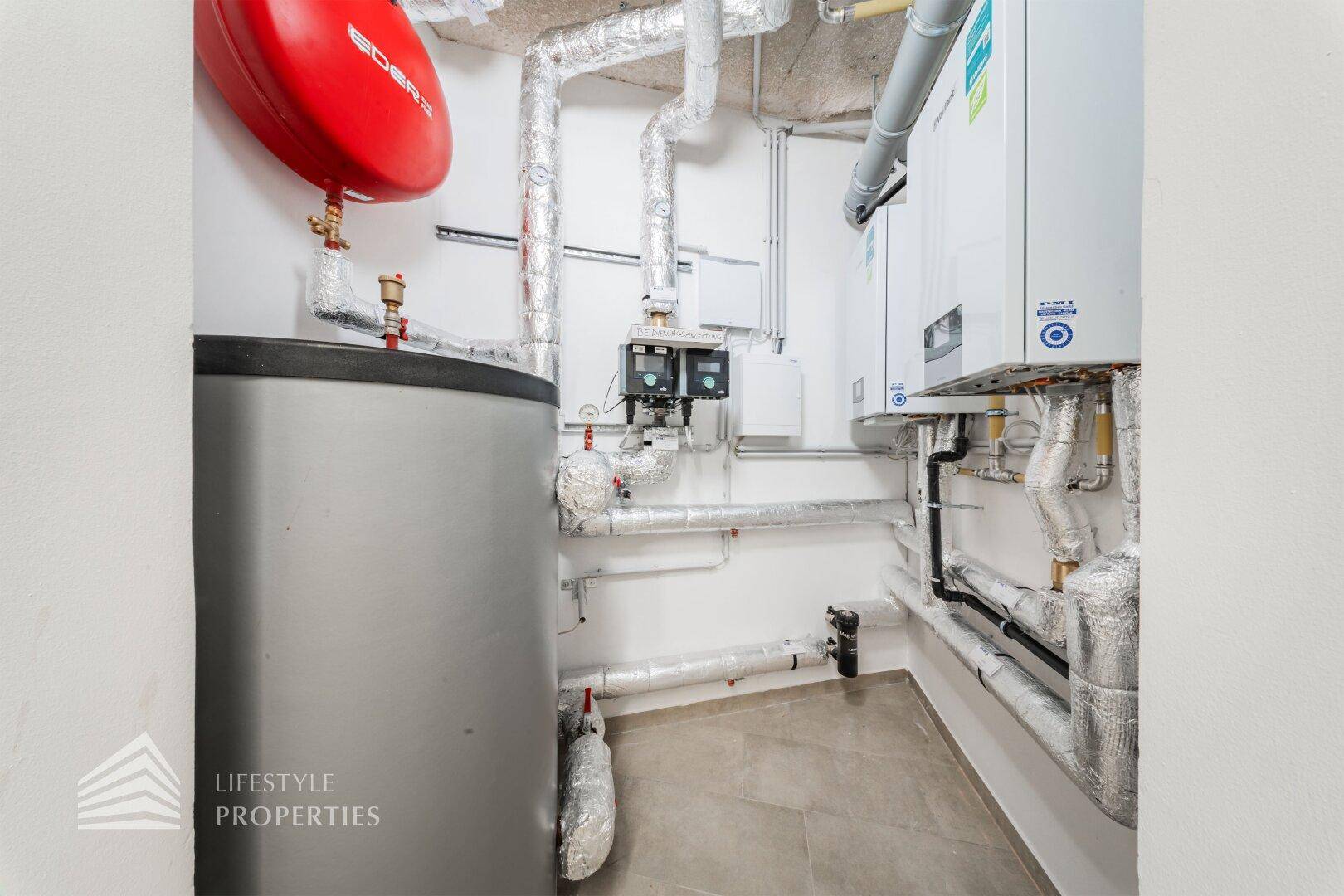 Technikraum mit Warmwasserspeicher, Heizungsanlage und isolierten Rohren an den Wänden.
