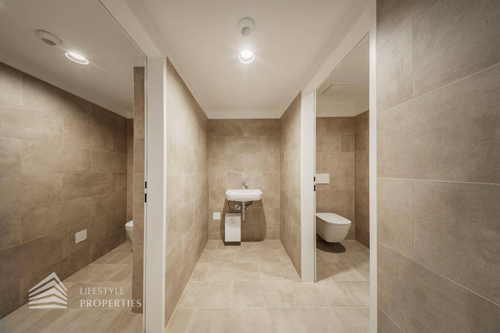 Moderner Sanitärbereich mit beigefarbenen Fliesen, Waschbecken und mehreren Toilettenkabinen.