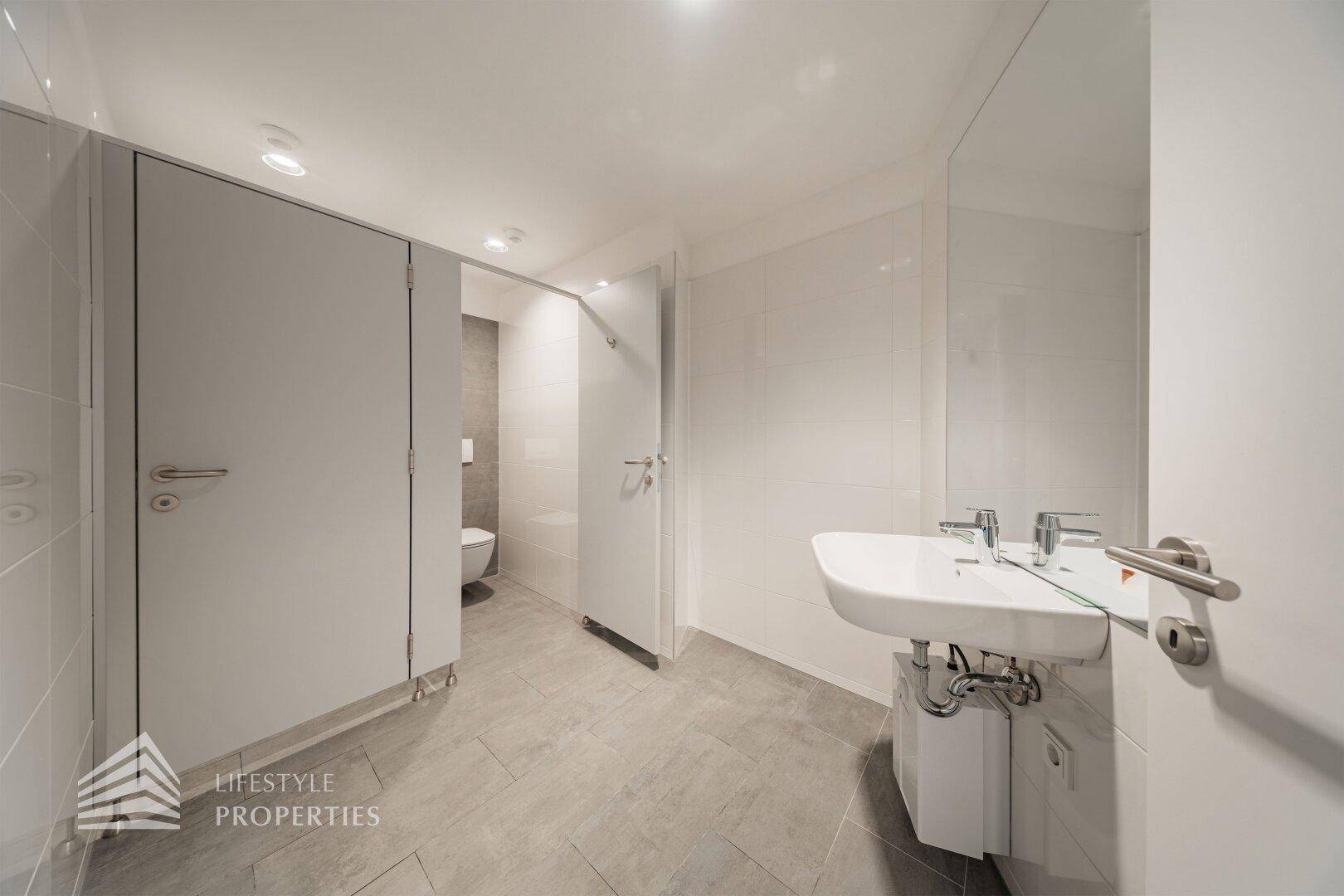 Modernes Badezimmer mit mehreren Toilettenkabinen, weißem Waschbecken und hellgrauen Fliesen.