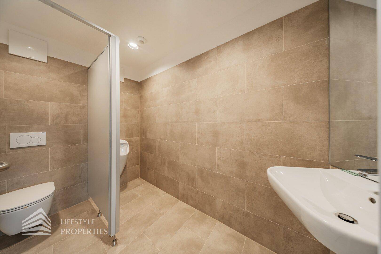 Modernes Badezimmer mit braunen Fliesen, Waschbecken und einer Toilettenkabine mit Urinal.