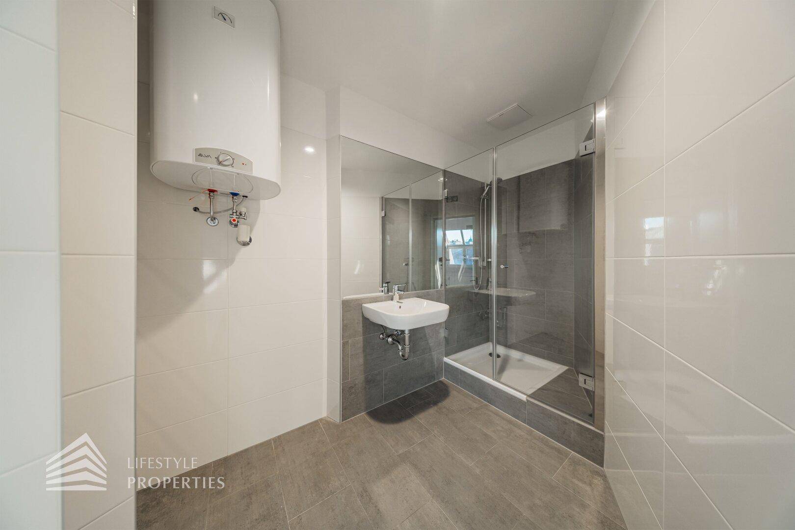 Modernes Badezimmer mit Duschkabine, Waschbecken und einem Warmwasserboiler, alles in hellen Farben gehalten.
