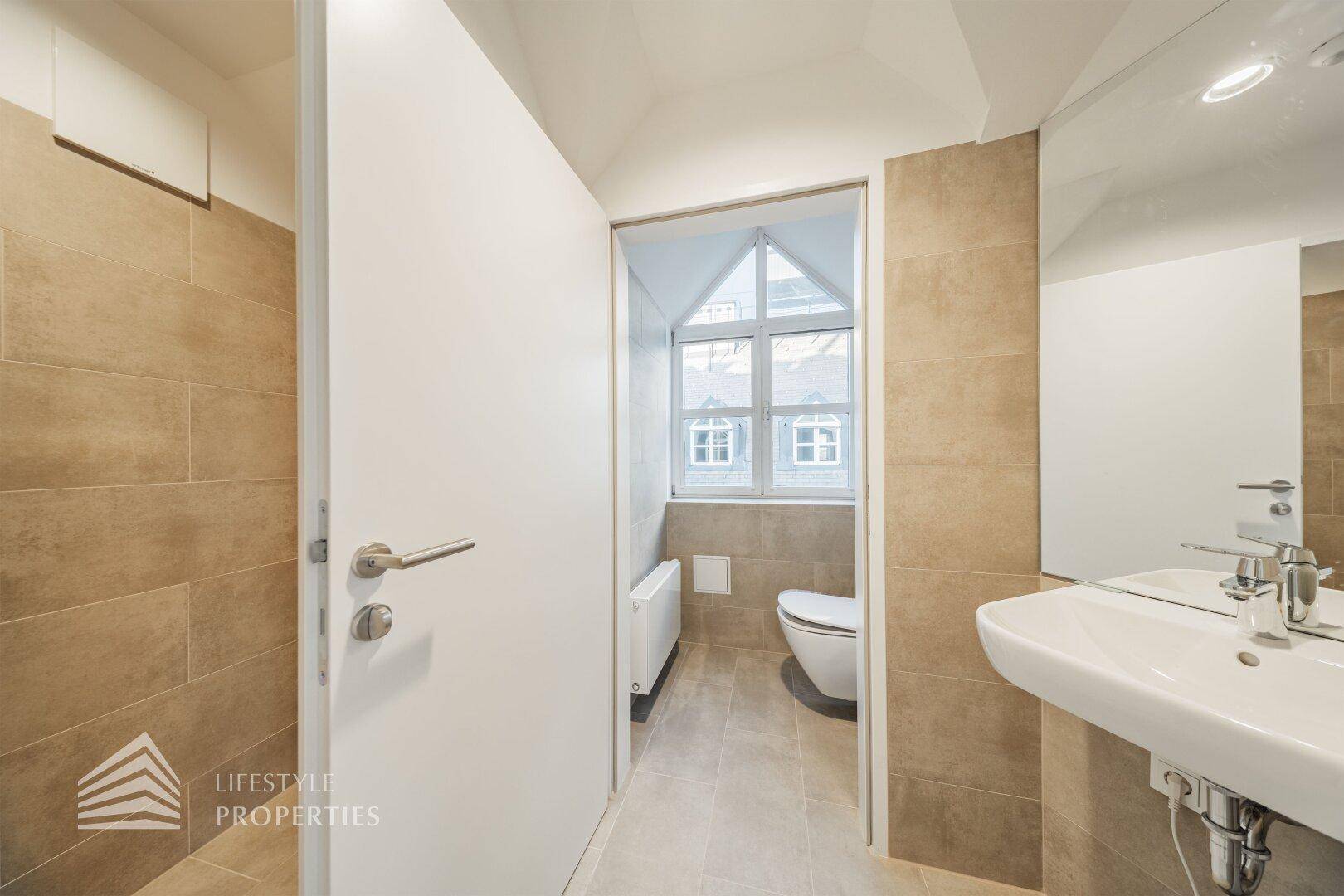 Separate Toilette mit beigefarbenen Fliesen, wandmontiertem WC und einem Fenster für natürliche Belüftung.