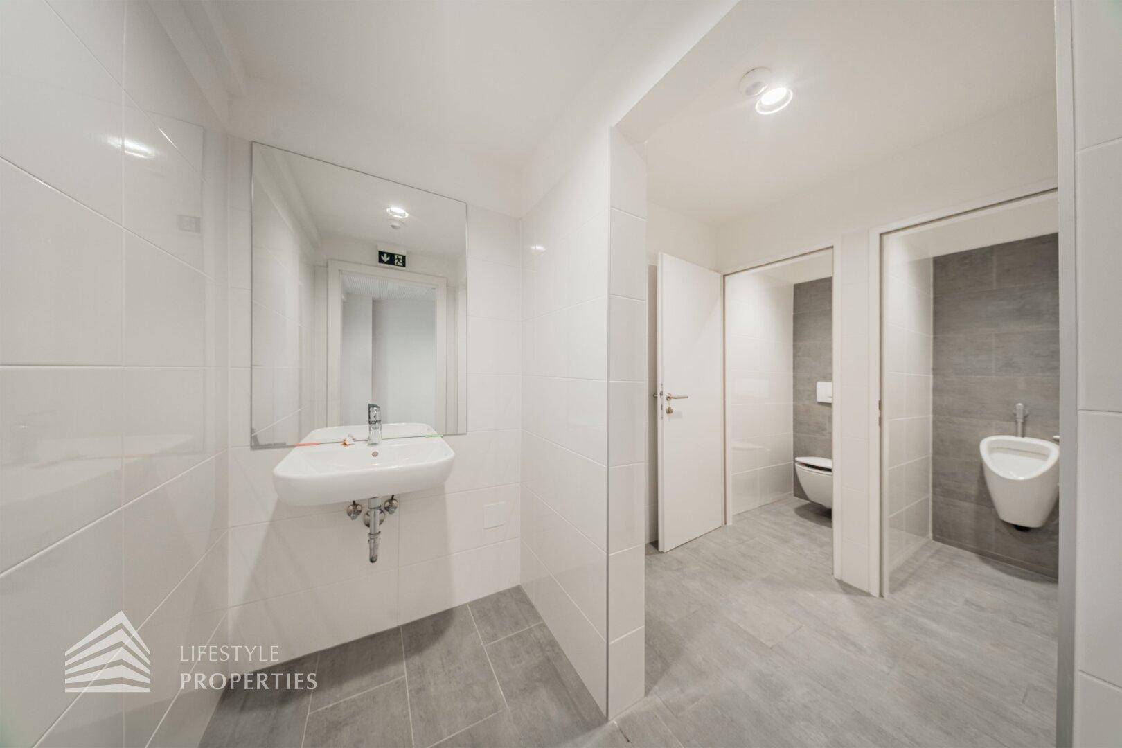Modernes Badezimmer mit Waschbecken, Spiegel, weißen Fliesen und separaten Toilettenkabinen.