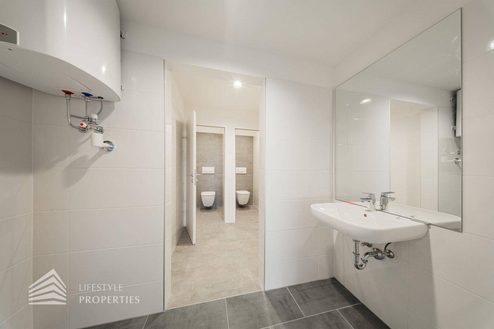 Badezimmer mit Boiler, Waschbecken, Spiegel und Zugang zu mehreren Toilettenkabinen.