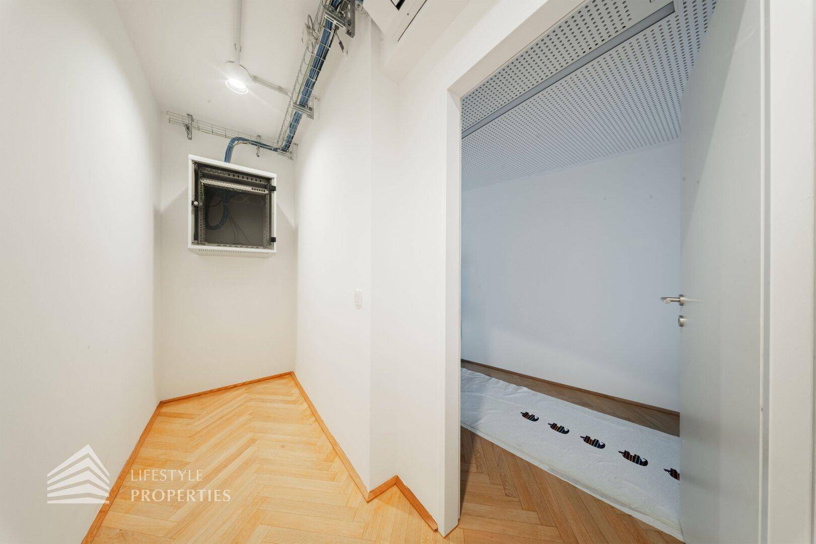 Kleiner Technikraum mit sichtbaren Leitungen und Sicherungskasten, angrenzend an einen Bürobereich.
