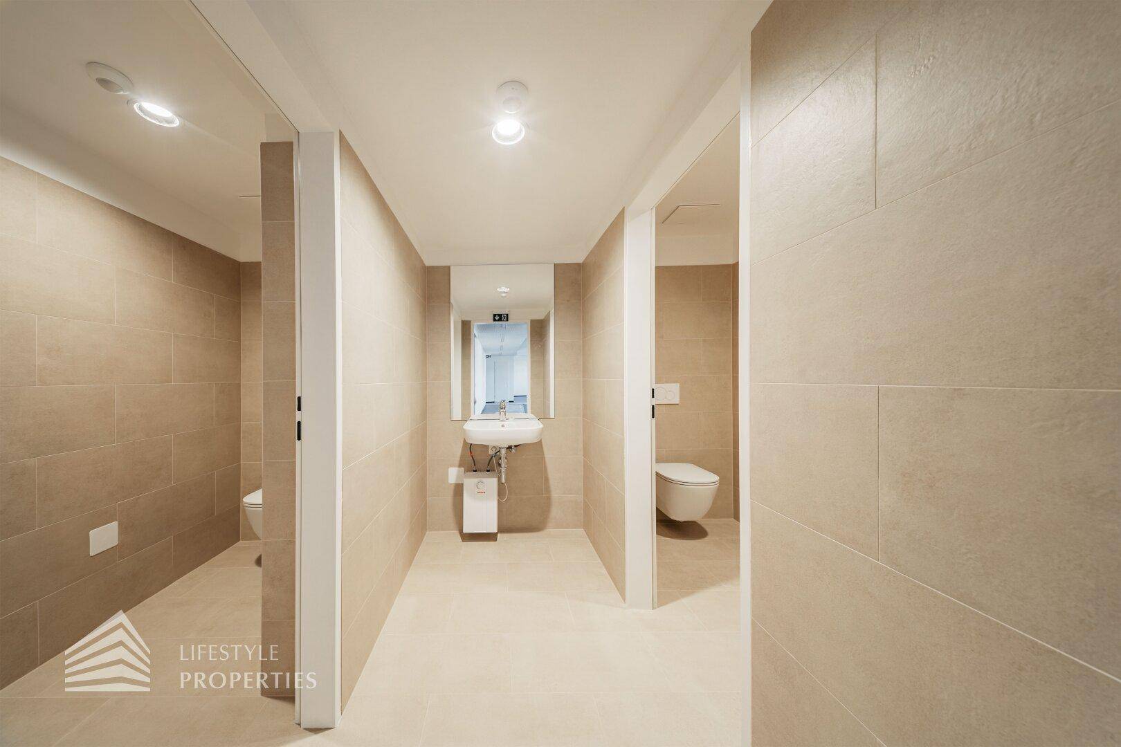Moderner Flur mit beigefarbenen Fliesen und Zugang zu mehreren Toiletten, ausgestattet mit Waschbecken und Spiegel.