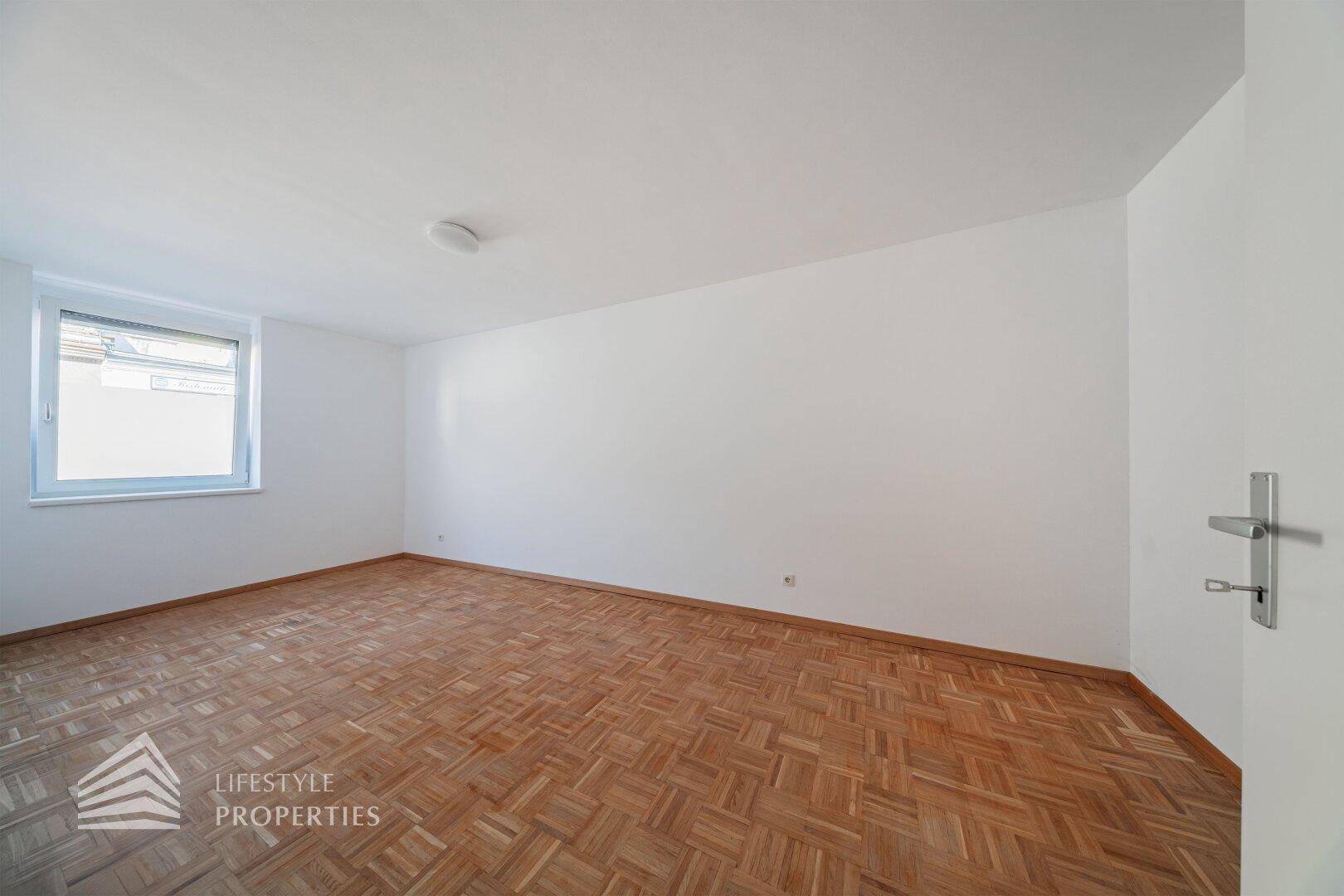 Helles Zimmer mit Parkettboden und einem Fenster, das für eine angenehme Atmosphäre sorgt.