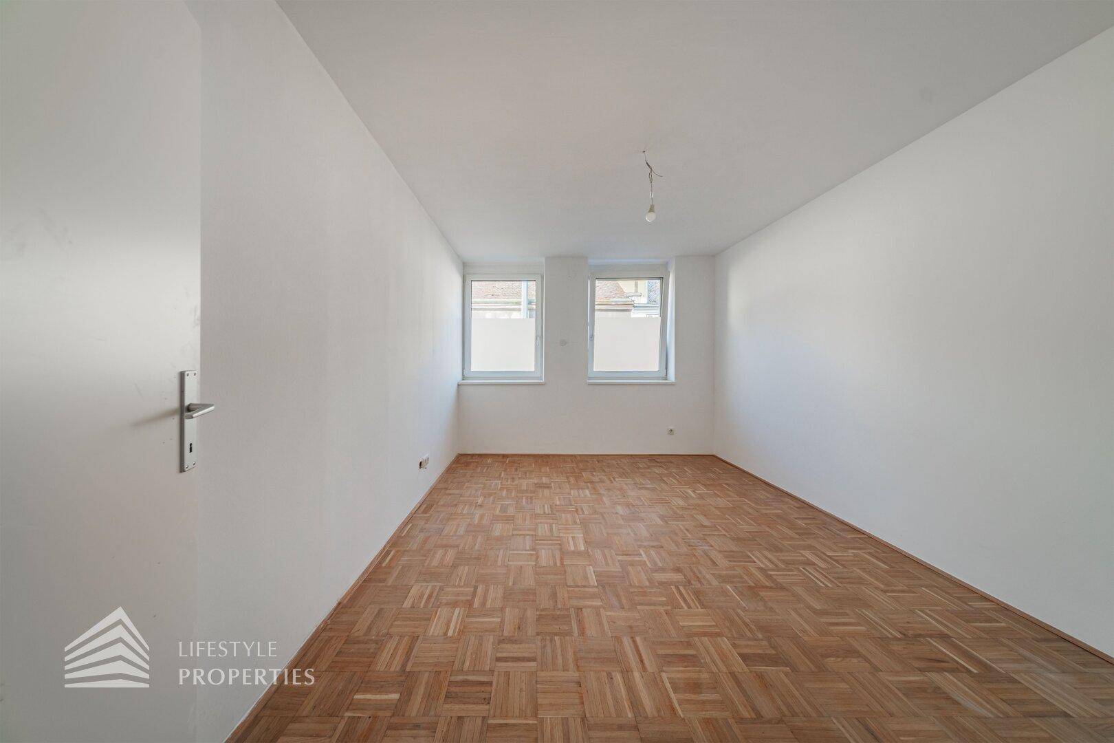 Großes Zimmer mit zwei Fenstern und Parkettboden, ideal als Schlaf- oder Arbeitszimmer.