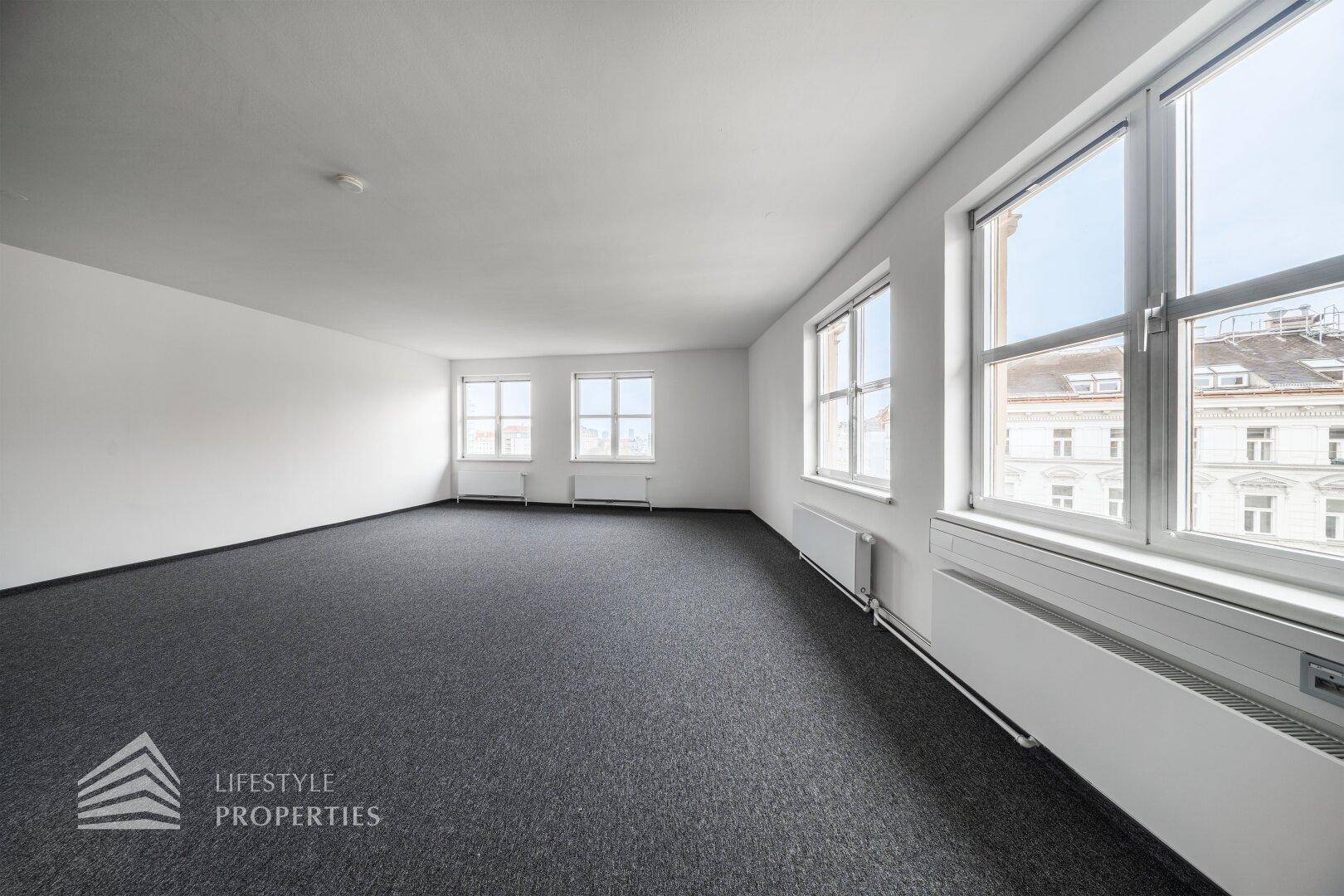 Heller Büroraum mit Teppichboden, großen Fenstern und Heizkörpern, bietet viel Tageslicht.
