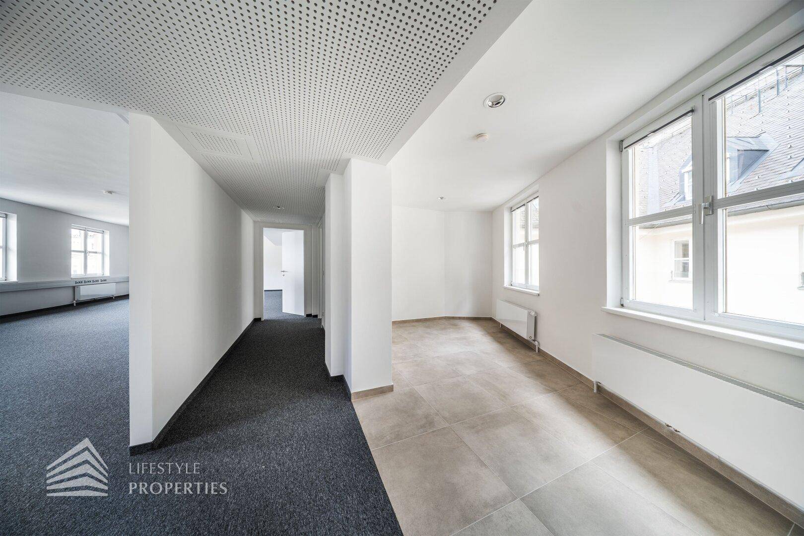 Übergangsbereich mit Teppichboden und Fliesen, perforierter Decke und natürlichem Lichteinfall durch Fenster.