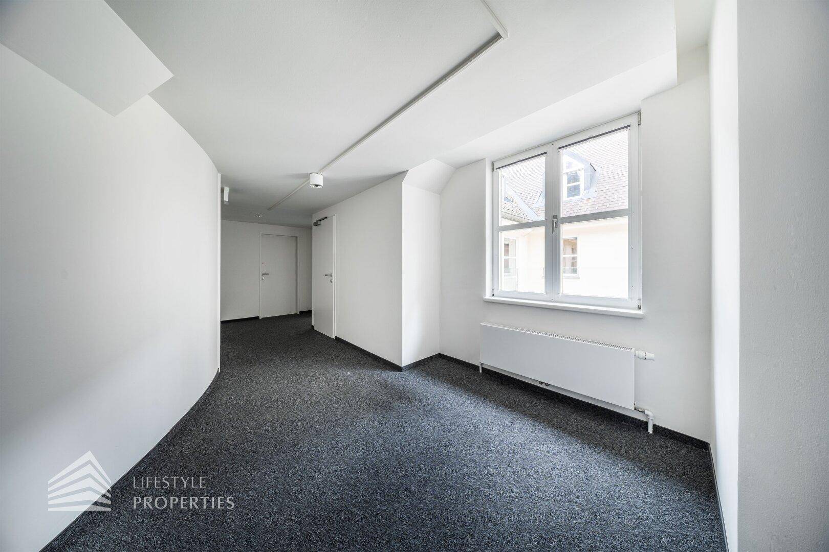 Heller Flur mit Teppichboden, weißen Wänden und einem Fenster, das Tageslicht in den Bereich bringt.