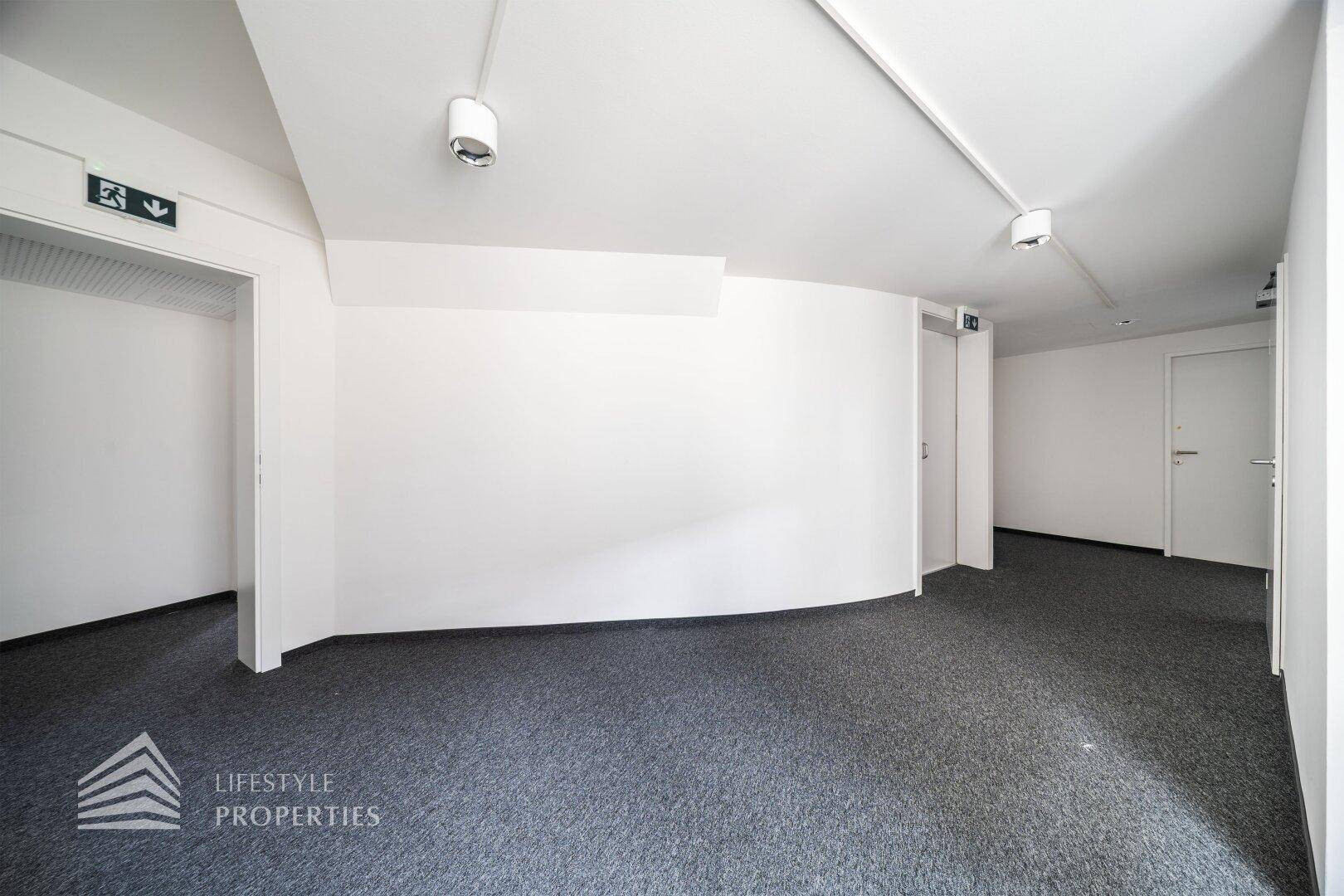Langer Flur mit Teppichboden, weißen Wänden und Notausgangsschildern, ideal für Büronutzung.