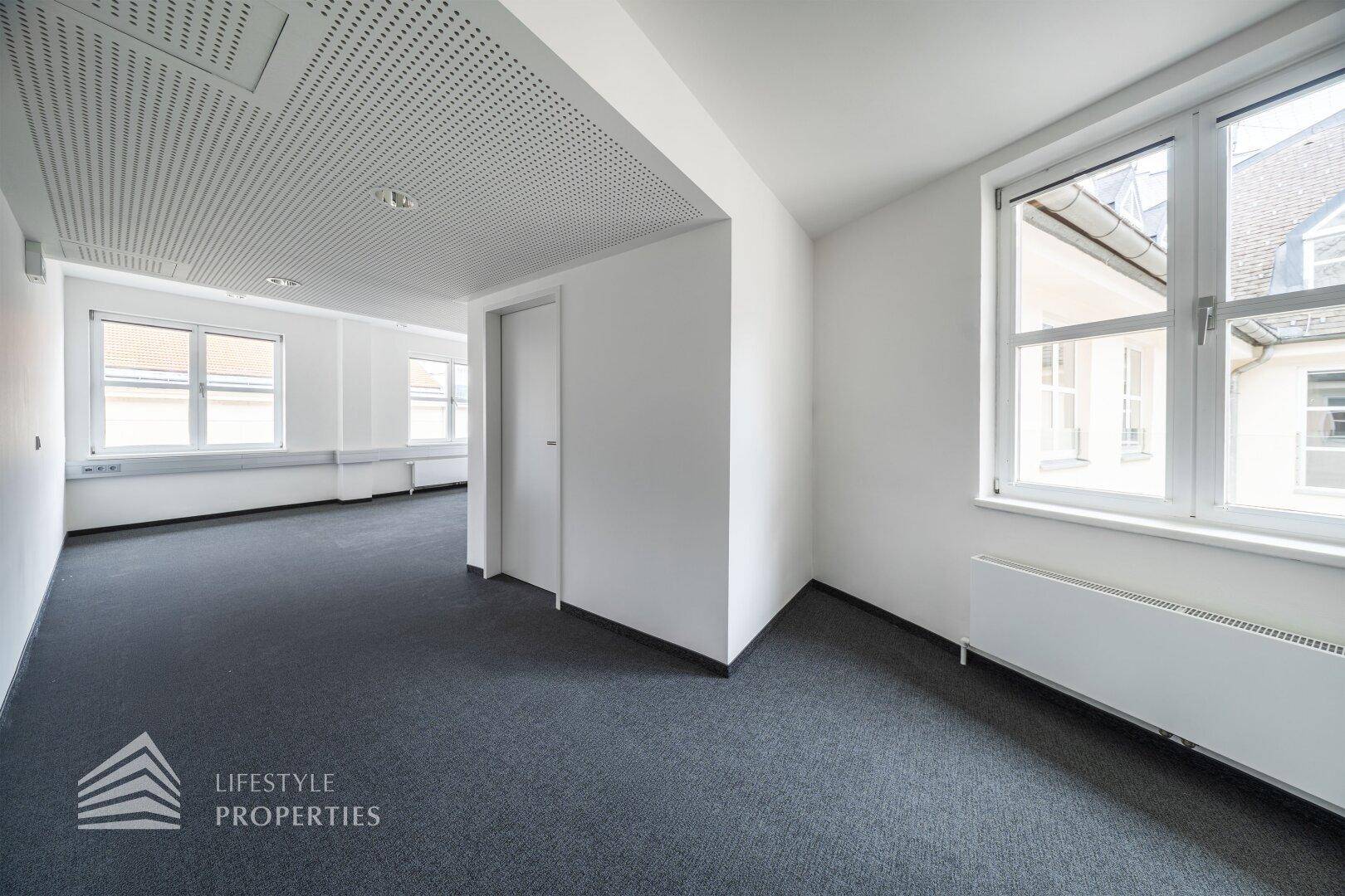 Offener Bürobereich mit Teppichboden, perforierter Decke und Fenstern, die den Raum erhellen.