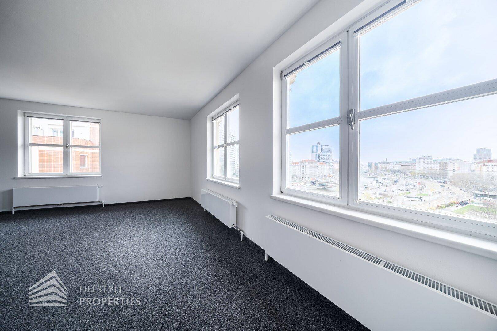 Heller Büroraum mit weitem Stadtblick durch große Fenster und grauem Teppichboden.