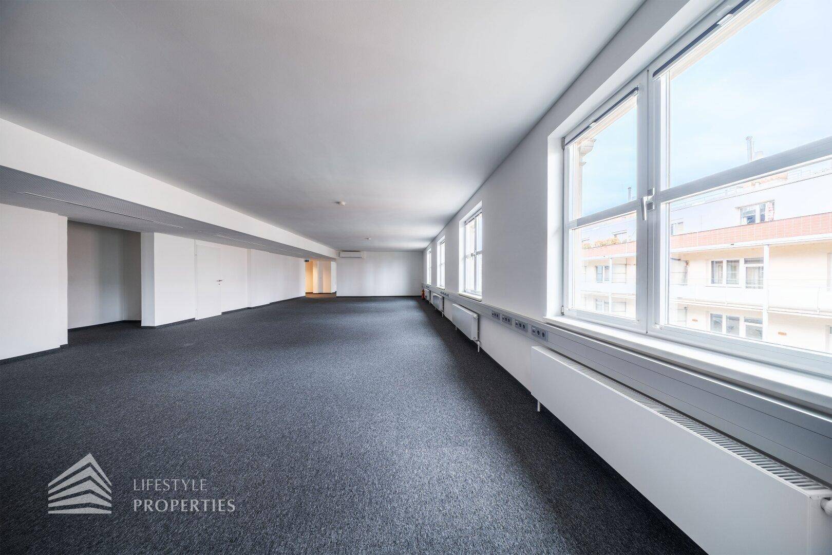 Heller Büroraum mit vielen Fenstern, die einen Blick auf die umliegenden Gebäude bieten.