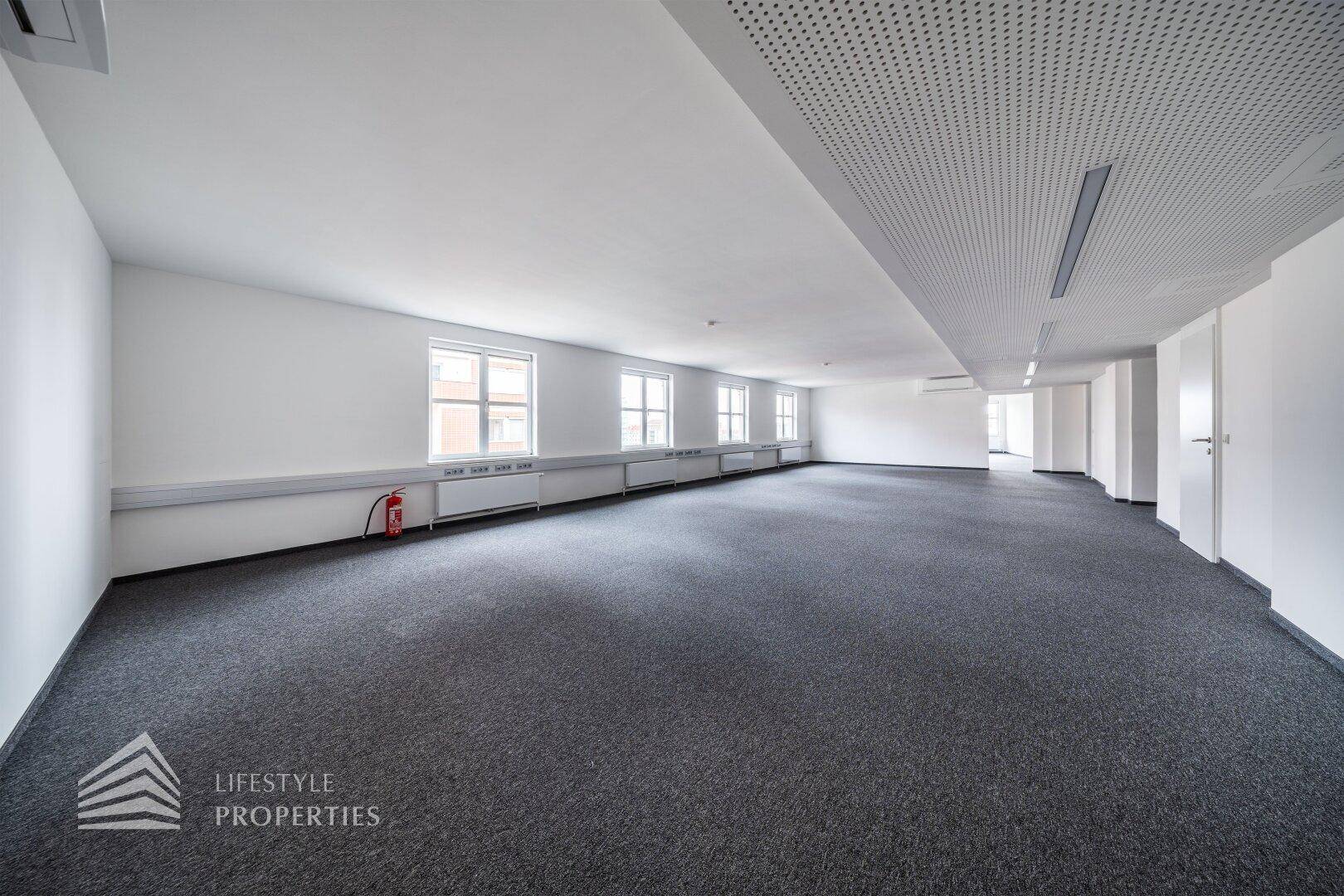 Großer, offener Büroraum mit Feuerlöscher, Teppichboden und perforierter Decke.