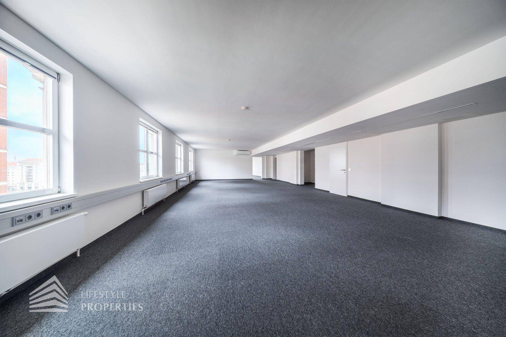 Langer Büroraum mit vielen Fenstern, die viel Licht hereinlassen, und moderner Deckenbeleuchtung.