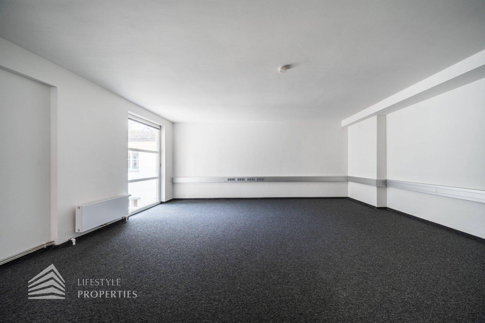 Leeres Büro mit grauem Teppichboden, weißen Wänden und einem Fenster, das den Raum mit Tageslicht versorgt.