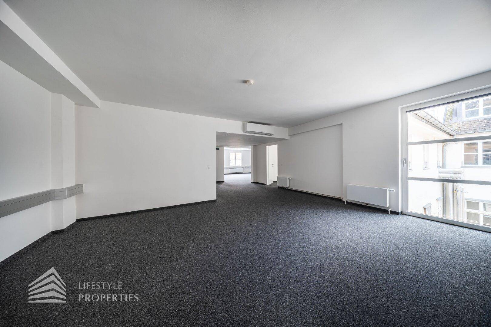 Geräumiges, leeres Büro mit grauem Teppichboden, weißen Wänden und einem großen Fenster mit Blick nach draußen.
