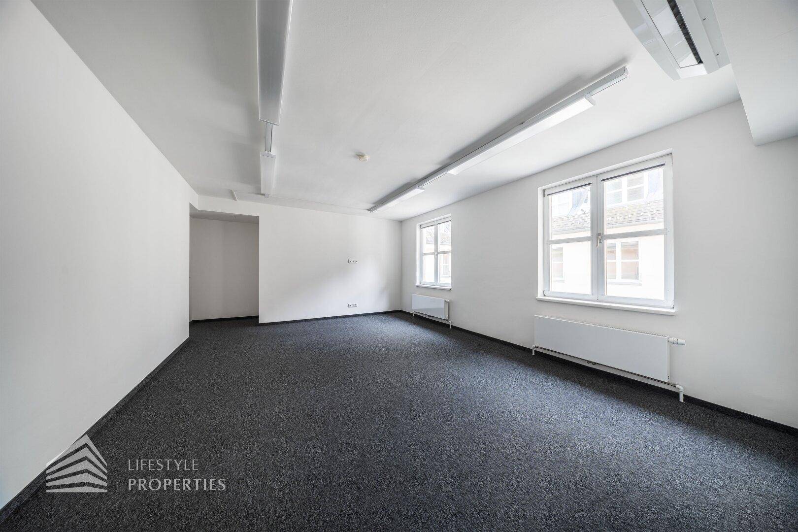 Kompaktes, leeres Büro mit grauem Teppichboden, weißen Wänden und zwei Fenstern für ausreichend Tageslicht.