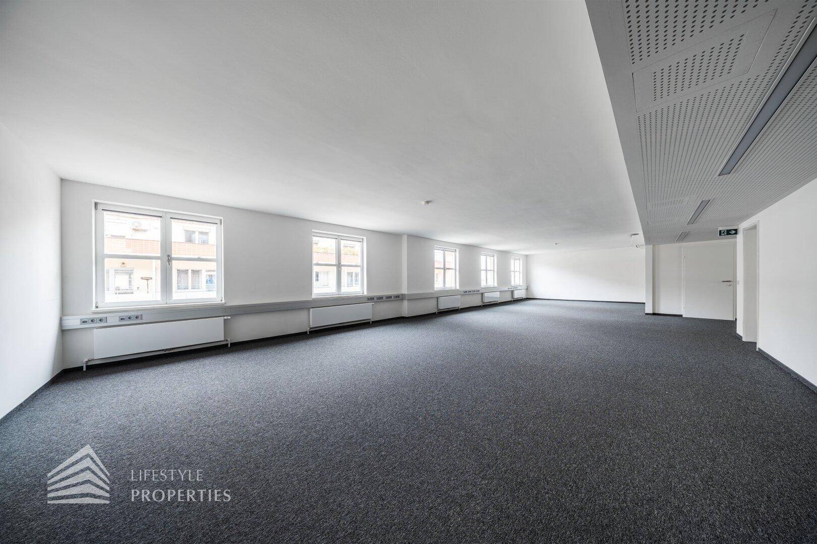 Großes, leeres Büro mit grauem Teppichboden, weißen Wänden und einer Reihe von Fenstern, die den Raum erhellen.