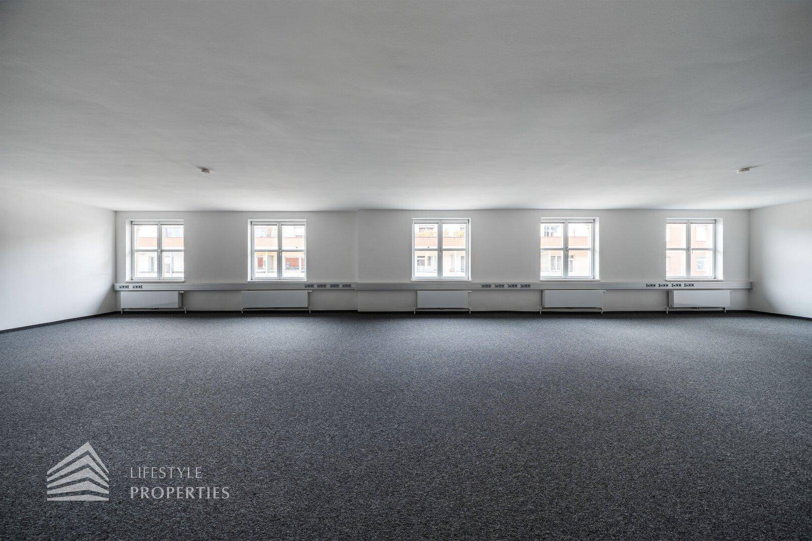 Langes, leeres Büro mit grauem Teppichboden, weißen Wänden und einer Fensterfront für viel Tageslicht.