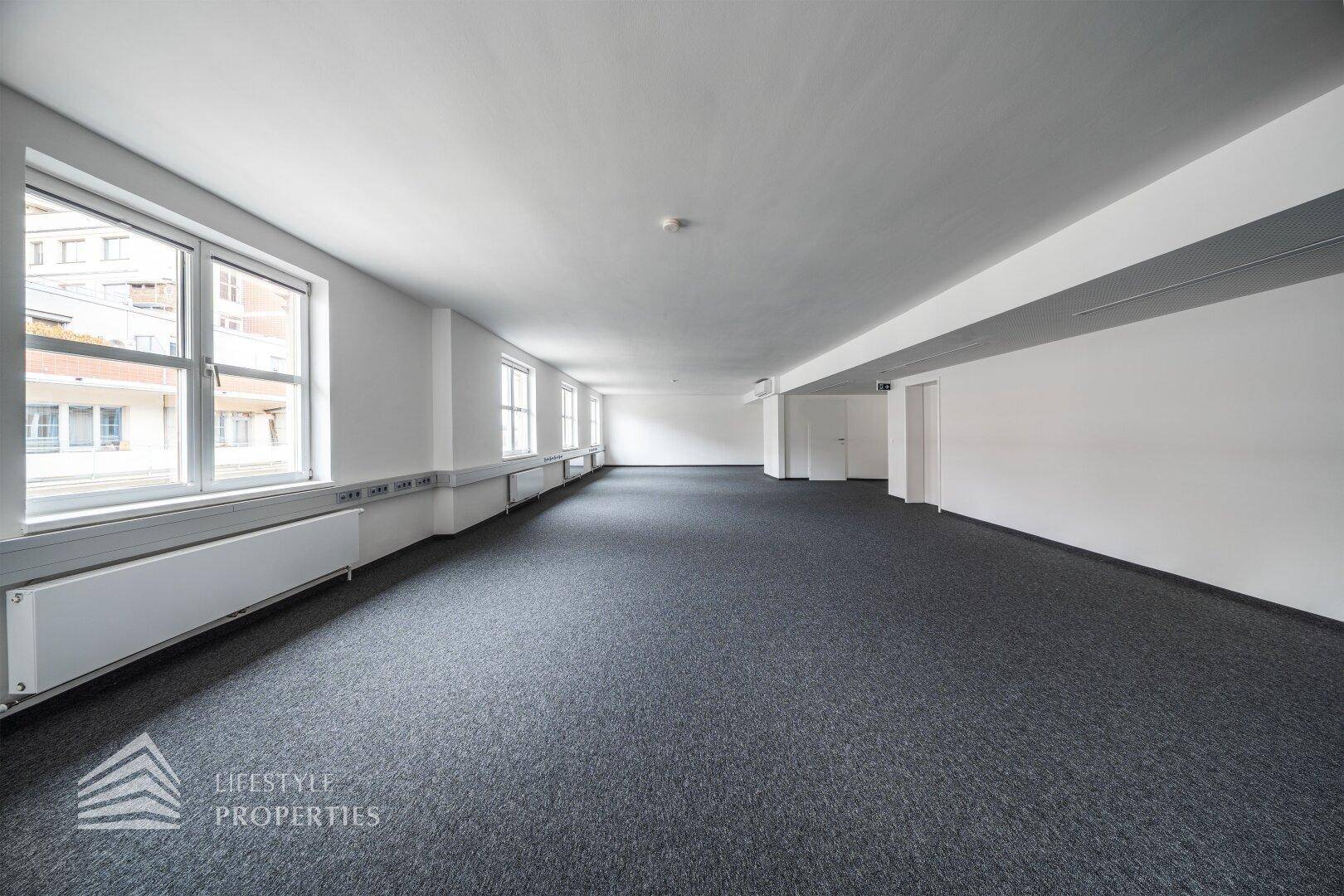Helles und geräumiges Büro mit grauem Teppichboden, weißen Wänden und mehreren Fenstern mit Stadtblick.
