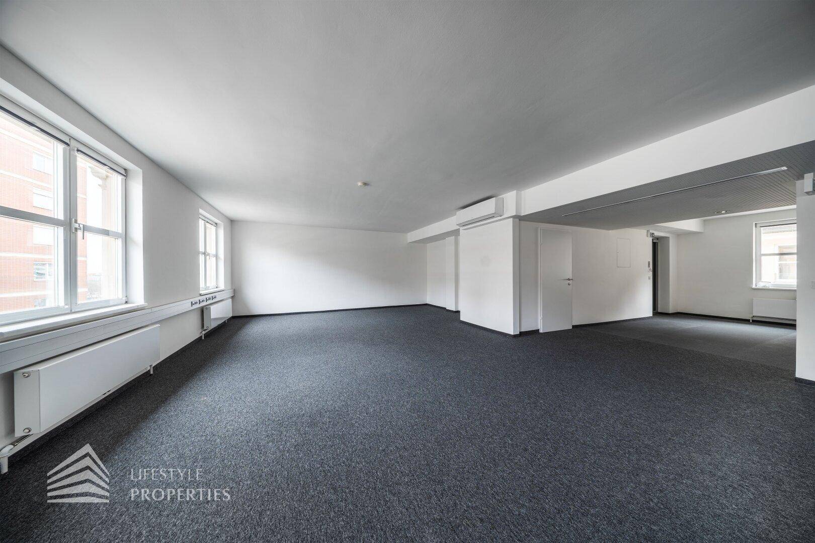 Weitläufiges Büro mit grauem Teppichboden, weißen Wänden und einer Reihe von Fenstern mit Stadtblick.