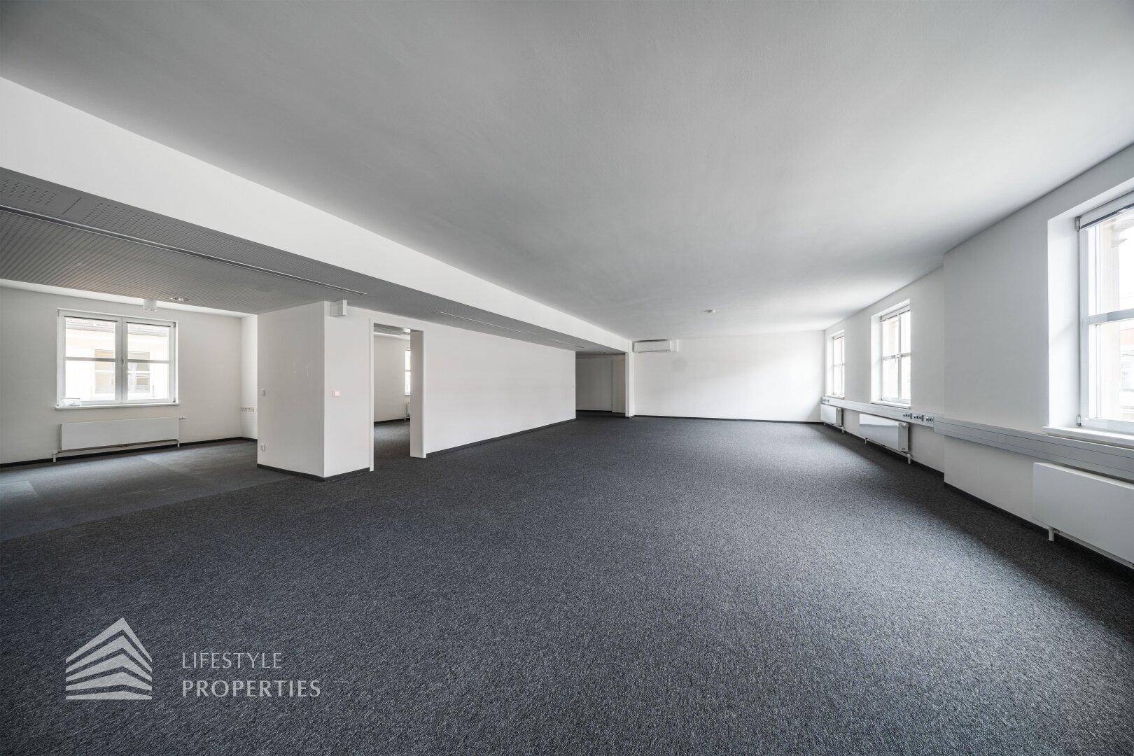 Großes, offenes Büro mit grauem Teppichboden, weißen Wänden und vielen Fenstern, die viel Licht hereinlassen.