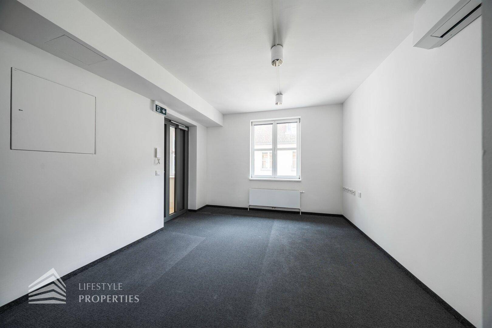 Helles, leeres Büro mit grauem Teppichboden, weiß gestrichenen Wänden und einem Fenster für Tageslicht.